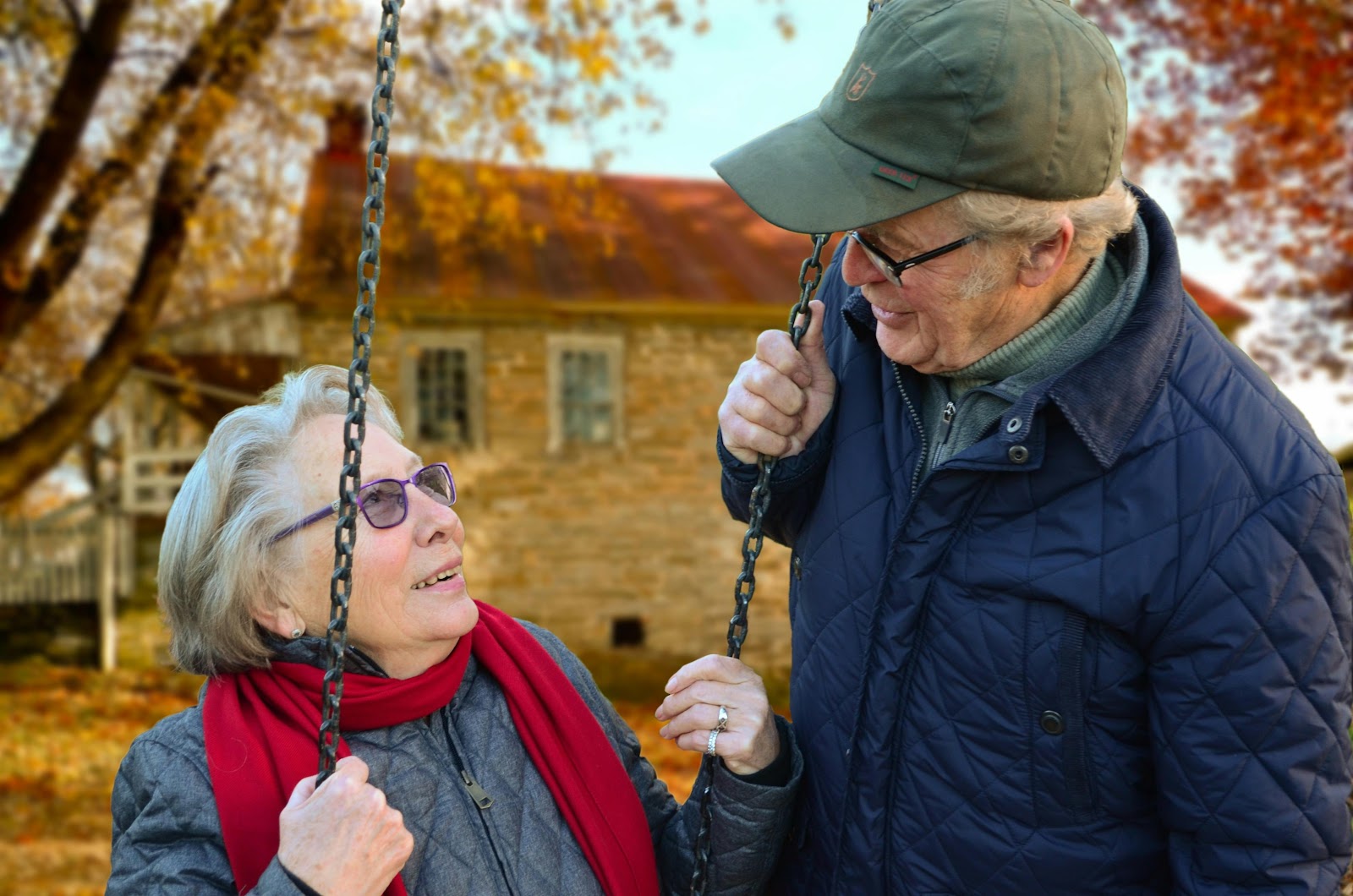  An elderly couple smiling and talking outdoors on a sunny autumn day, standing near a swing, with a rustic house and colorful fall leaves in the background.