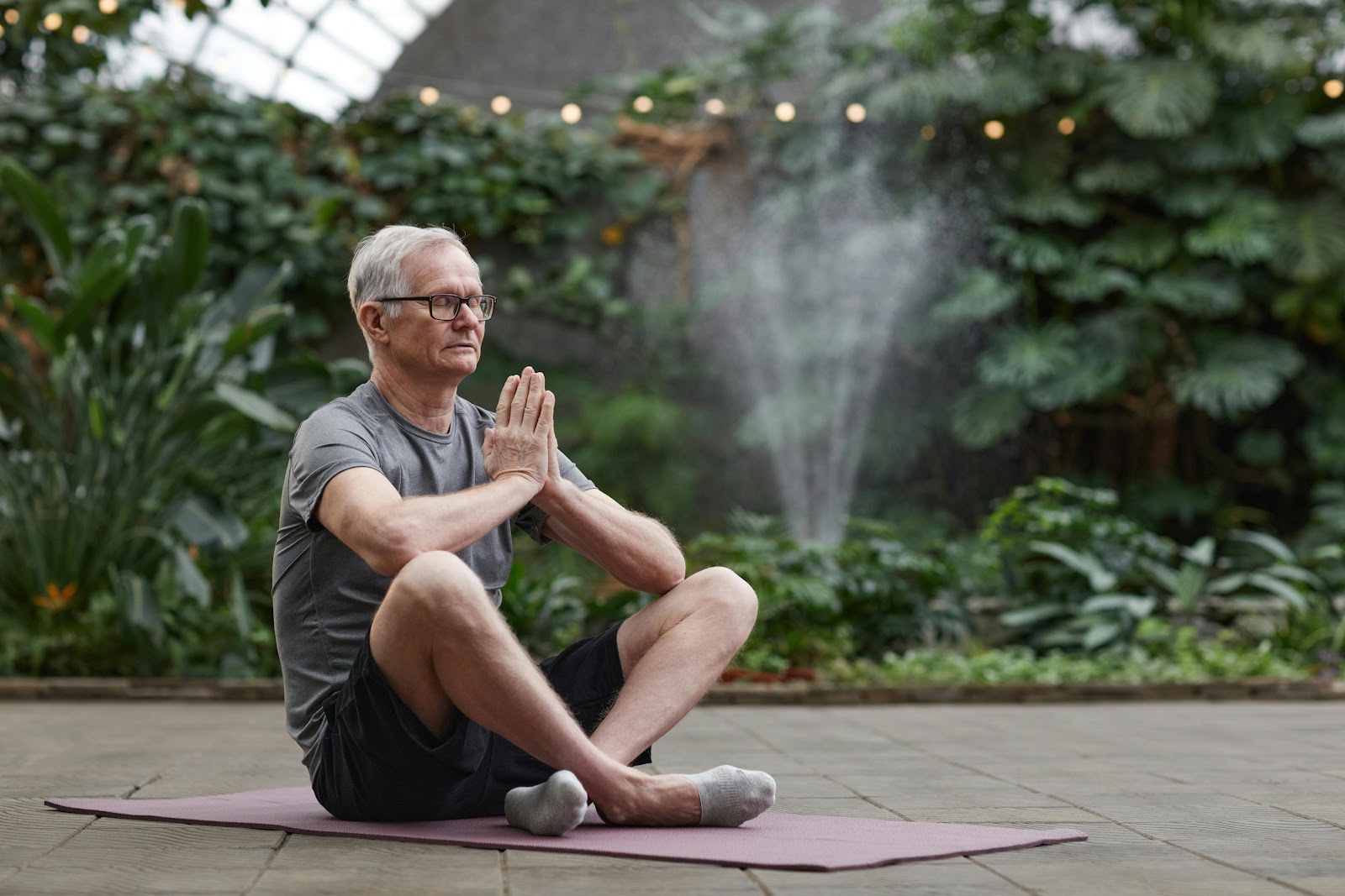 An older man with gray hair and glasses sits cross-legged on a yoga mat in a peaceful greenhouse, practicing meditation with his hands in a prayer position. 