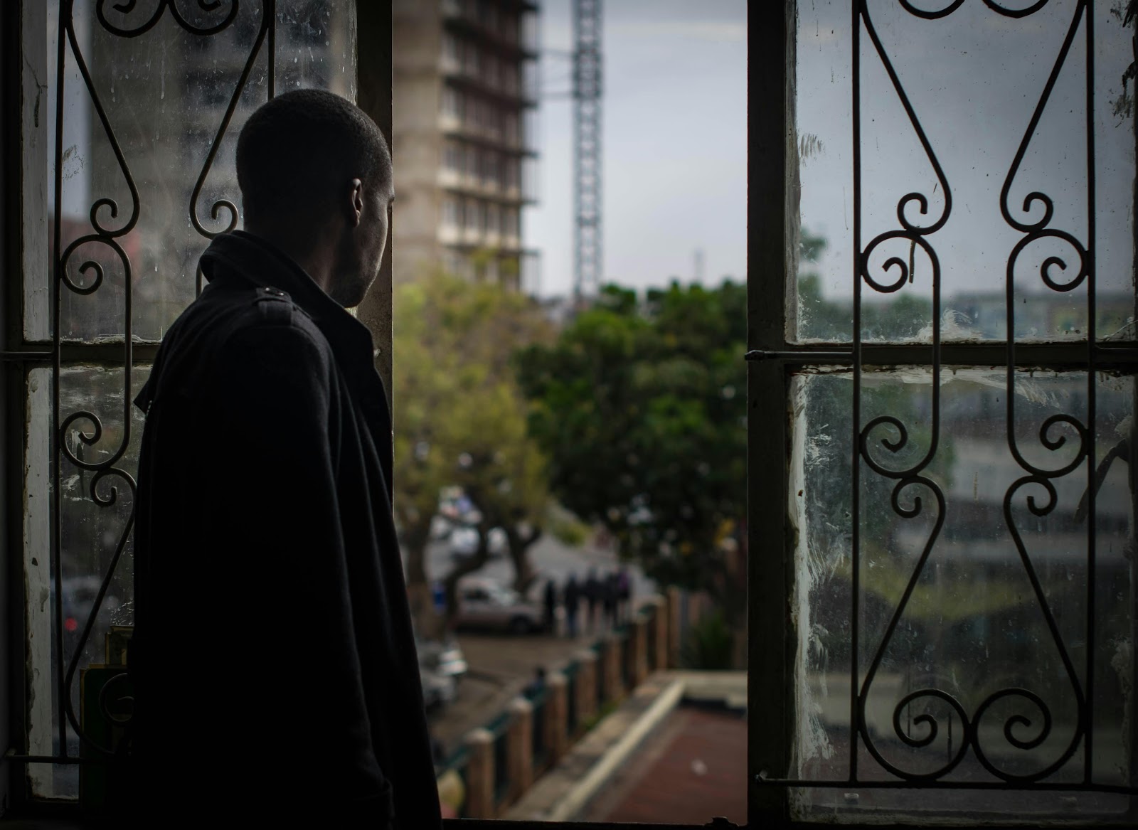 A man standing by a window, looking out at a city street, with trees and buildings visible in the background.