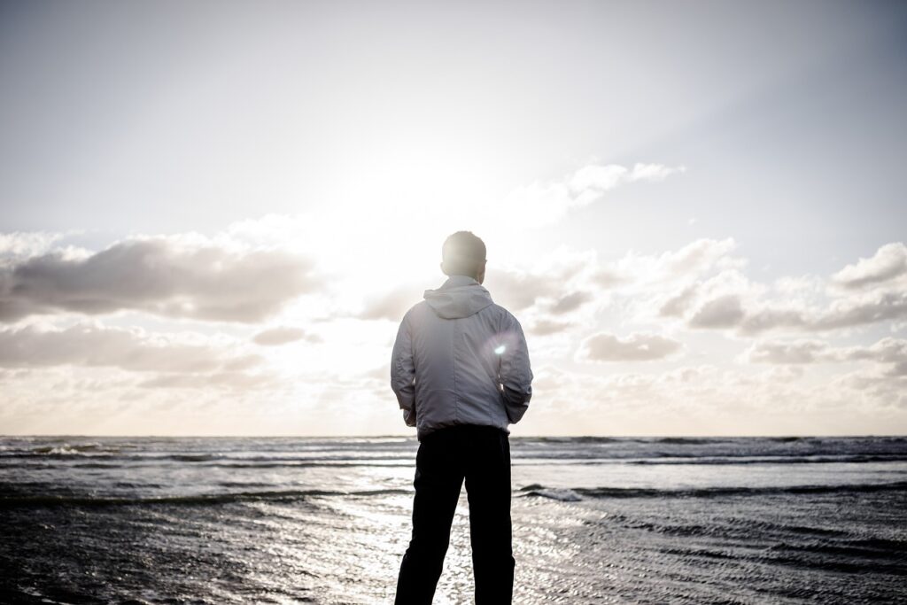 A man in a white jacket stands on the shore, facing the ocean at sunrise or sunset, with bright sunlight breaking through the clouds.