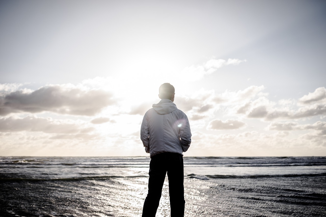 A man in a white jacket stands on the shore, facing the ocean at sunrise or sunset, with bright sunlight breaking through the clouds.